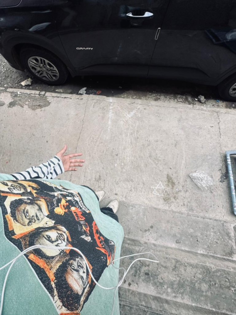 "A top-down view of a dark grey car parked next to a textured concrete sidewalk. Below it, a person's upper body is visible, wearing a green patterned shirt, lying on the sidewalk. They are gesturing with their left hand and holding an object, possibly a phone with wires."